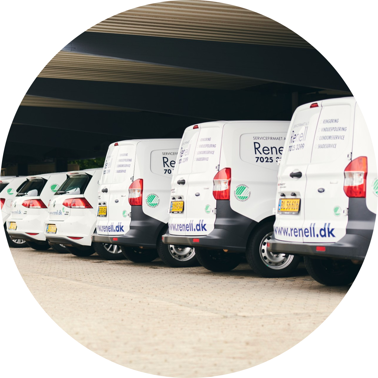 Række af hvide Renell servicebiler parkeret under et overdækket område med firmaets logo og hjemmeside synligt på bilerne.