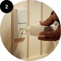 En hånd sætter et hvidt elektrisk stik i en stikkontakt på en beige væg. Rundt billede med sort cirkel og tallet 2.