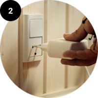 En hånd sætter et hvidt elektrisk stik i en stikkontakt på en beige væg. Rundt billede med sort cirkel og tallet 2.