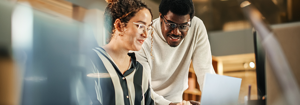 To personer med briller samarbejder foran en computerskærm i et moderne kontormiljø med varm belysning.