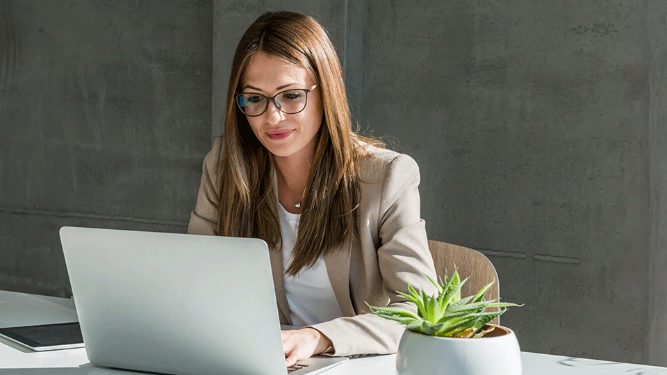 Person med briller og beige jakke arbejder på bærbar computer ved skrivebord med en lille grøn plante i hvid potte.