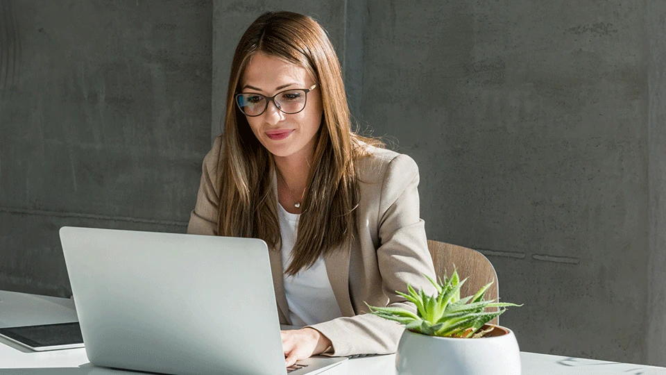 Person med briller og beige jakke arbejder på bærbar computer ved skrivebord med en lille grøn plante i hvid potte.
