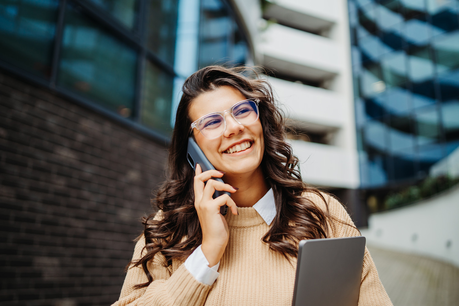Smilende person med briller taler i telefon foran moderne bygning, iført beige sweater og holder en bærbar computer.
