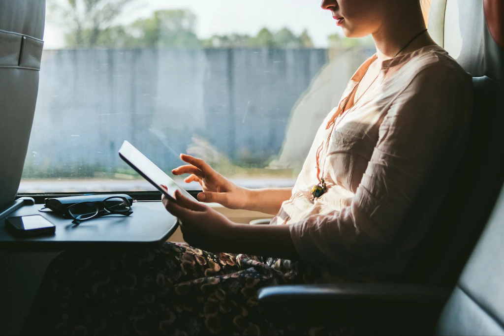 Person i lys skjorte bruger tablet i tog eller bus, med briller på bordet foran sig og udsigt gennem vinduet.