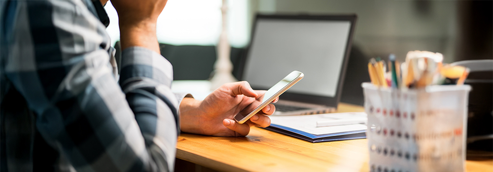 Person i ternet skjorte bruger smartphone ved træskrivebord med laptop og penneholder i baggrunden.